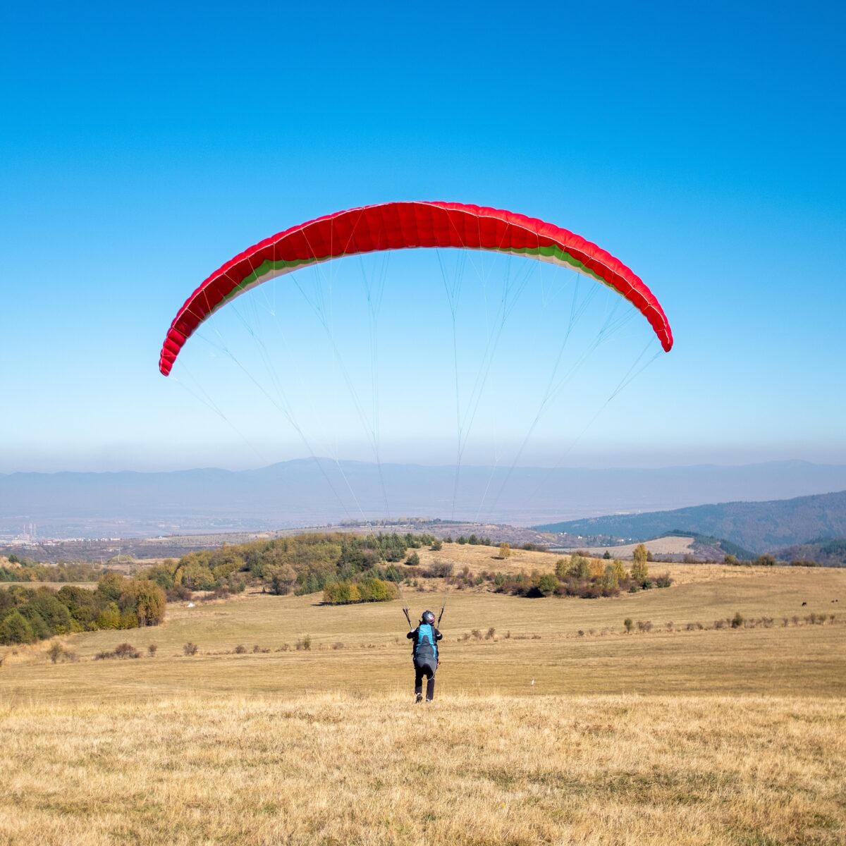 Vertical picture of a man flying with a red parachute surrounded by greenery under a blue sky Альбатрос-Ультра